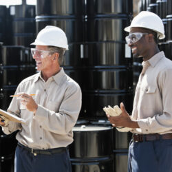 Senior man, 60s, with clipboard, taking inventory of steel barrels, with African American co-worker, 20s.